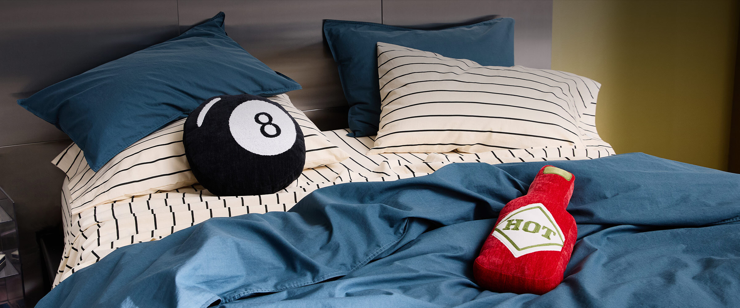Bed with a blue duvet cover, striped sheets, and two funky cushions