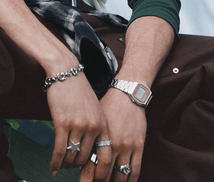 Gros plan sur des mains ornées de bagues, d'un bracelet argenté et d'une montre numérique, posées sur un pantalon brun.