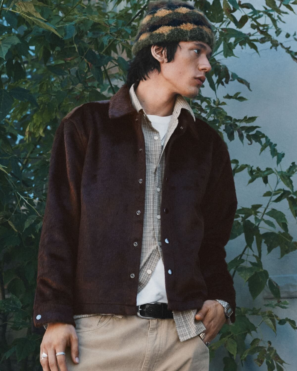 Homme devant un feuillage, vêtu d'une veste brun foncé, d'une chemise à carreaux, d'un pantalon beige et d'une tuque rayée.