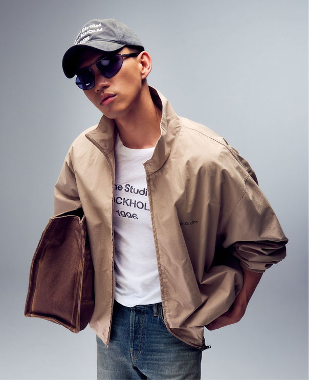 A young man wearing a cap, a beige jacket, a T-shirt with a logo, and jeans by Acne Studios.
