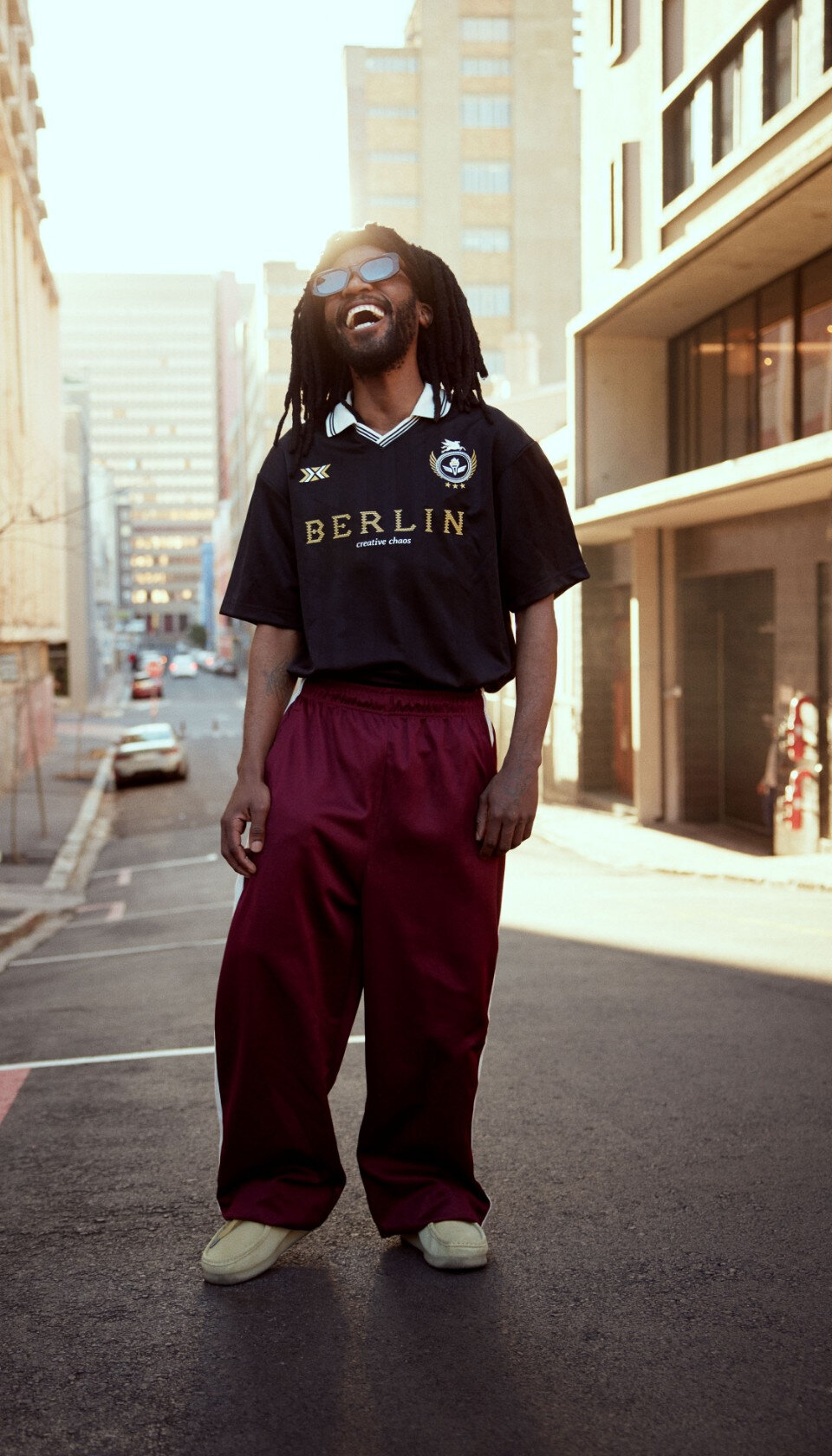 Homme dans une rue de ville ensoleillée, habillé dans un style sportif urbain, portant des lunettes de soleil et un large sourire radieux