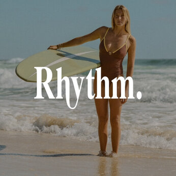 Une femme sur la plage avec une planche de surf porte un maillot une-pièce brun et jaune de marque Rhythm.