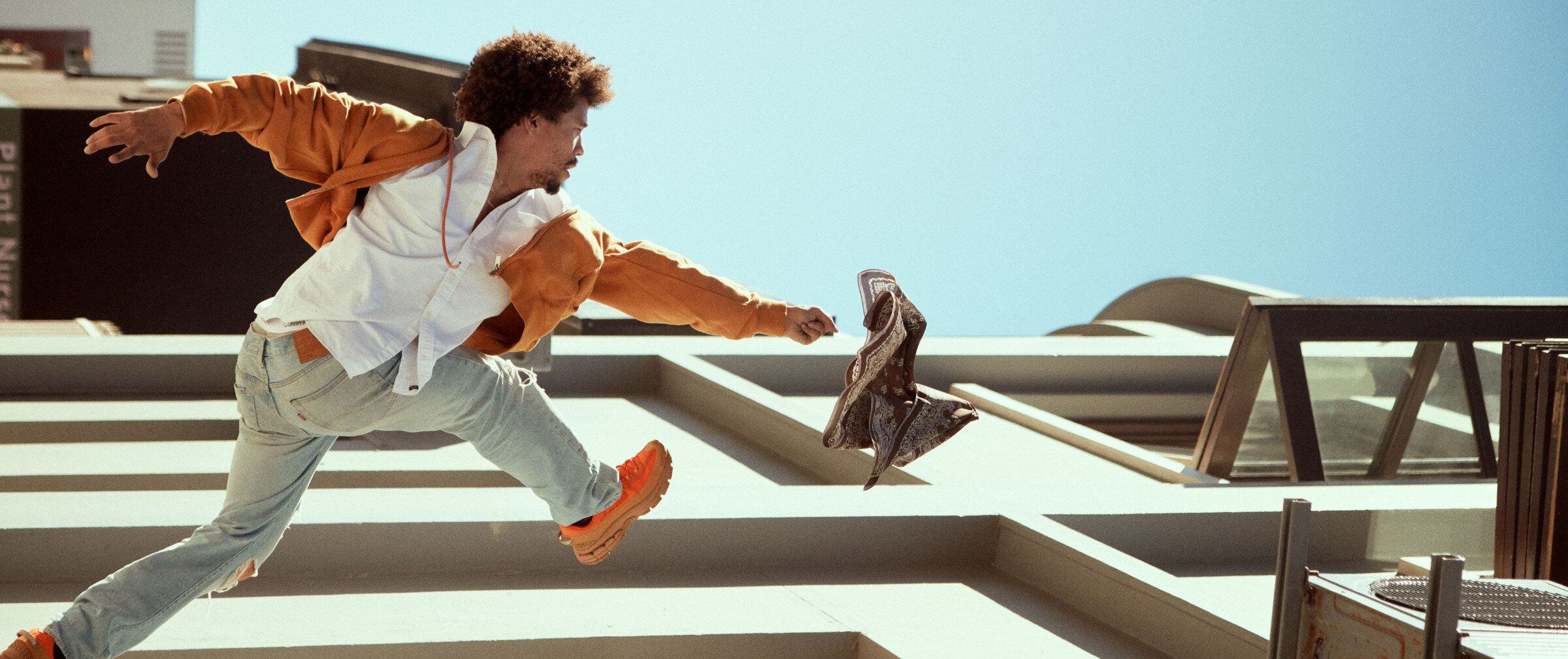 Jeune homme portant un jean, une chemise blanche, des sneakers et un kangourou zippé ouvert oranges, en train de courir sur le toit d'un édifice