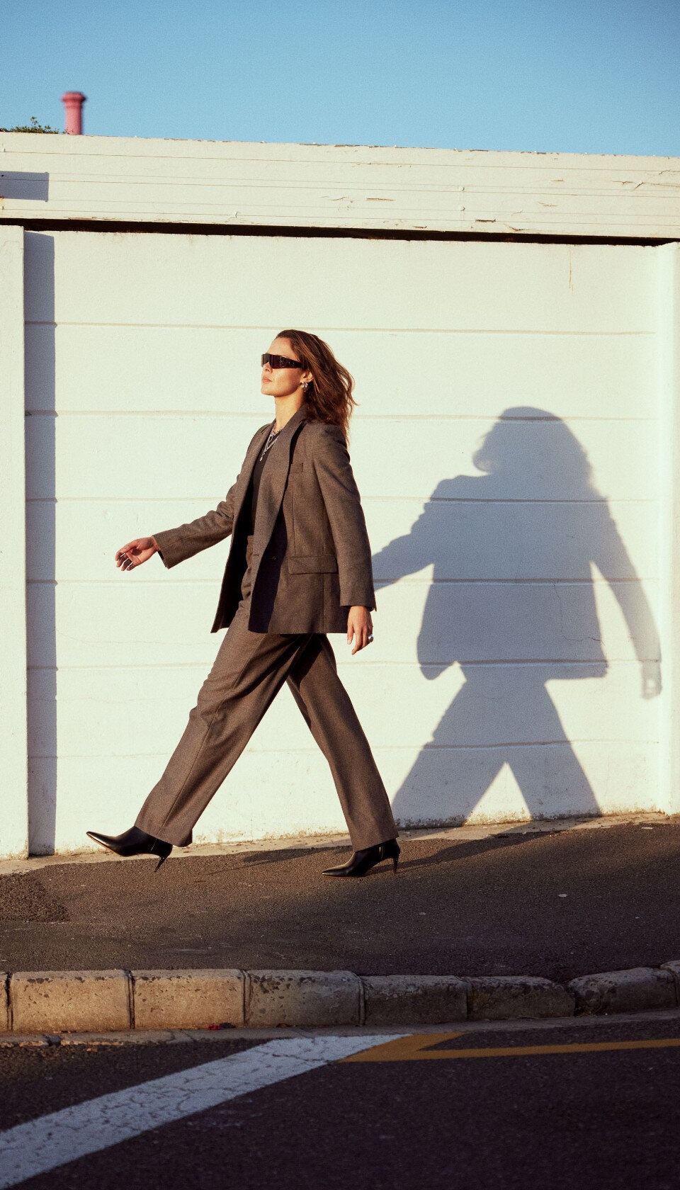 Professional-looking woman walking down a sunny street, wearing a brown pantsuit, sunglasses, and black leather heeled boots