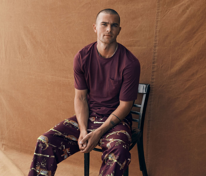 Man seated on a chair, wearing a burgundy T-shirt and matching pants patterned with big cats and foliage.