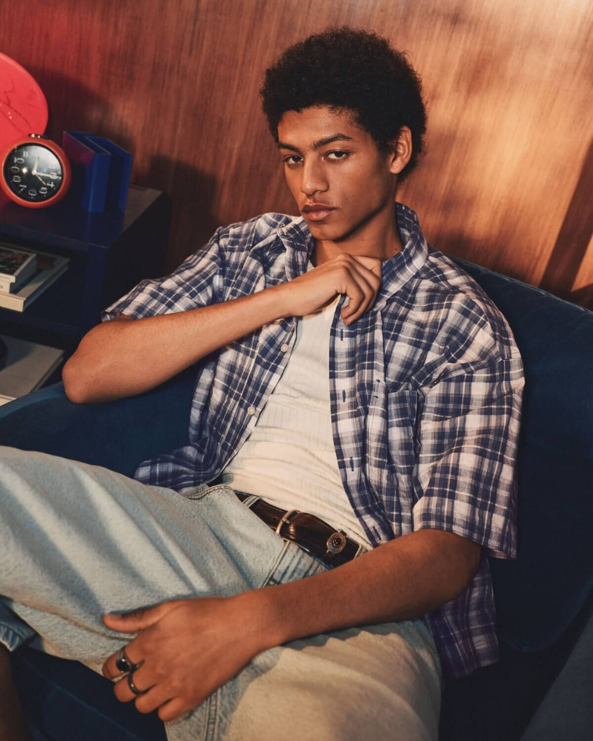 A young man wearing a short-sleeved plaid shirt left open over a white tank top