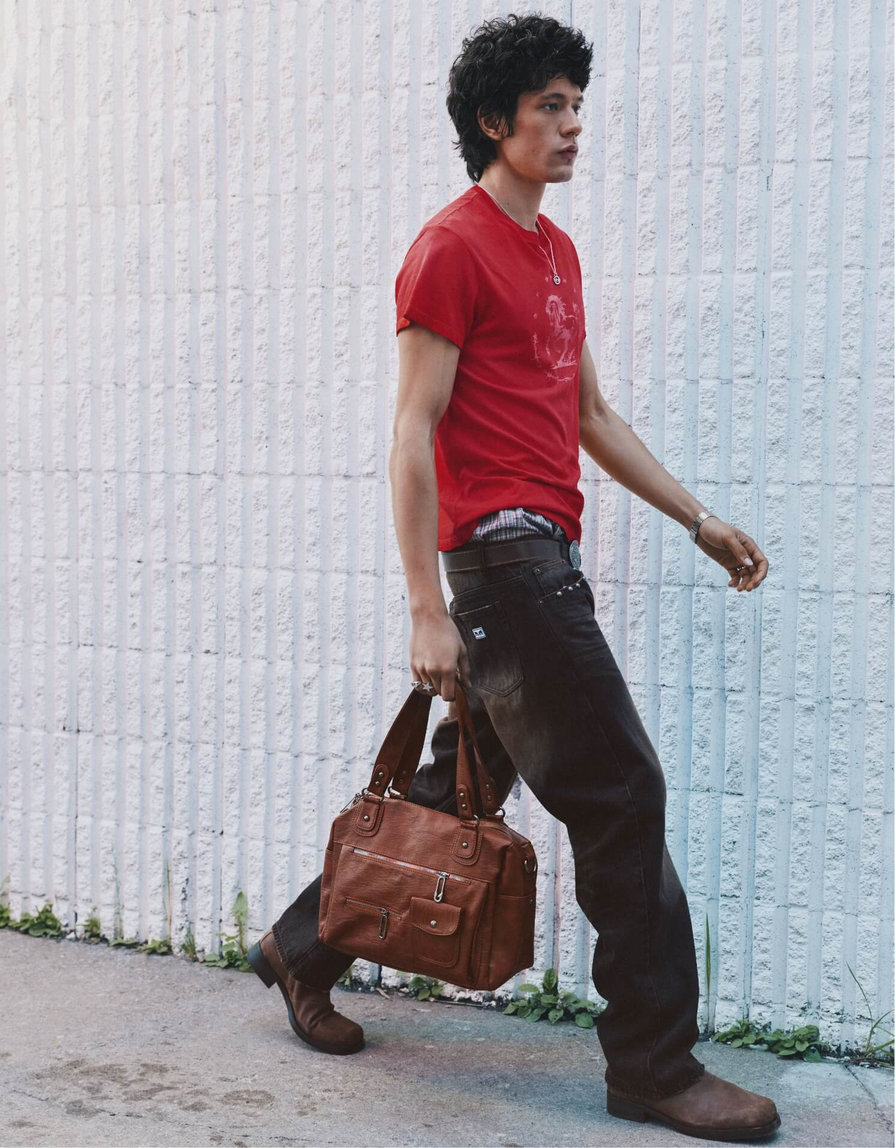 Man walking along a white wall with a brown bag, wearing a red T-shirt, dark jeans and brown boots.