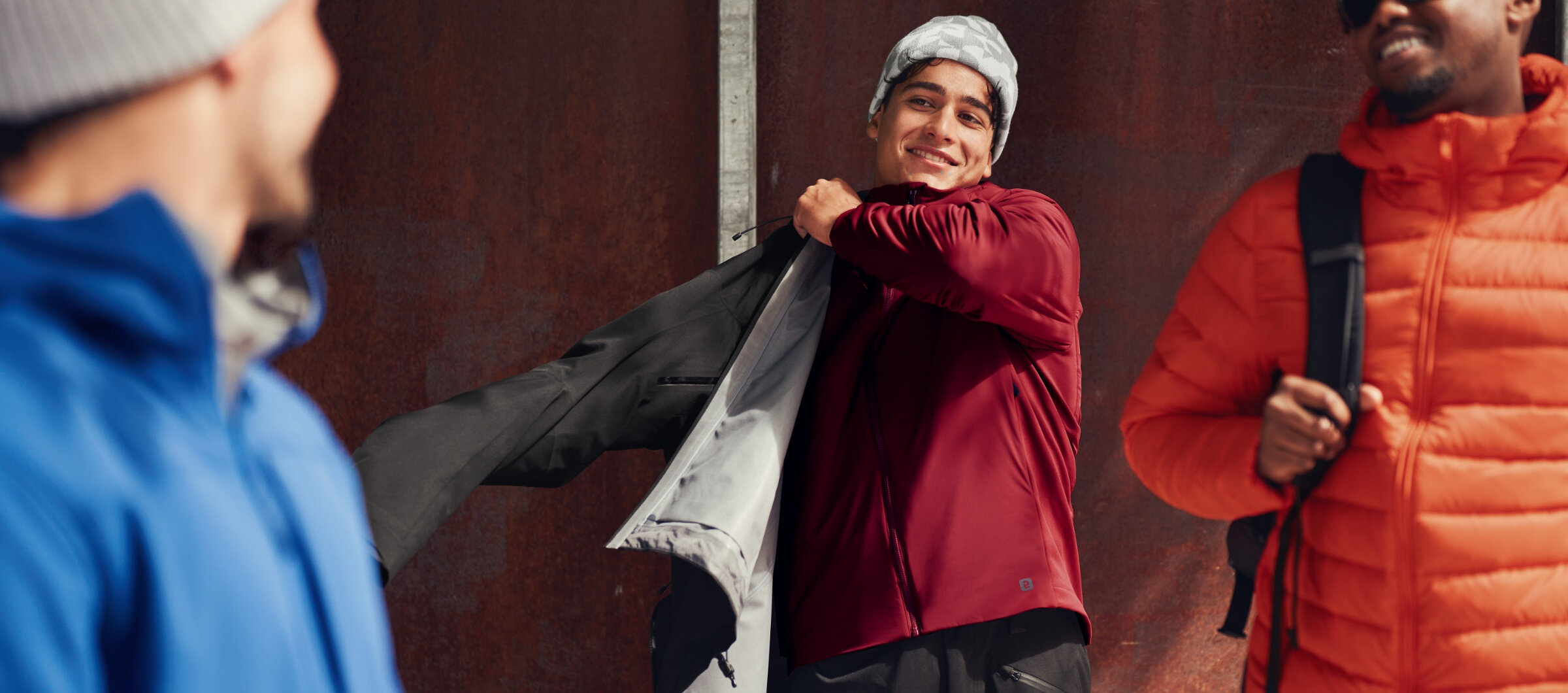 Homme enfilant une coquille imperméable noire par-dessus un manteau léger rouge avec une tuque grise à motif géométrique