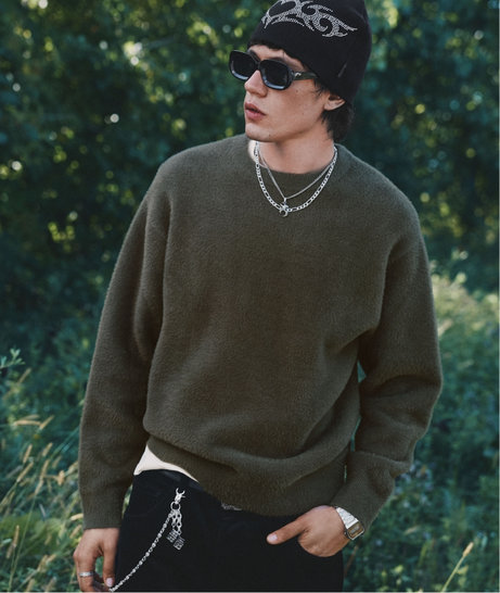 Man wearing a dark green sweater, black beanie, sunglasses and layered necklaces in a leafy setting.
