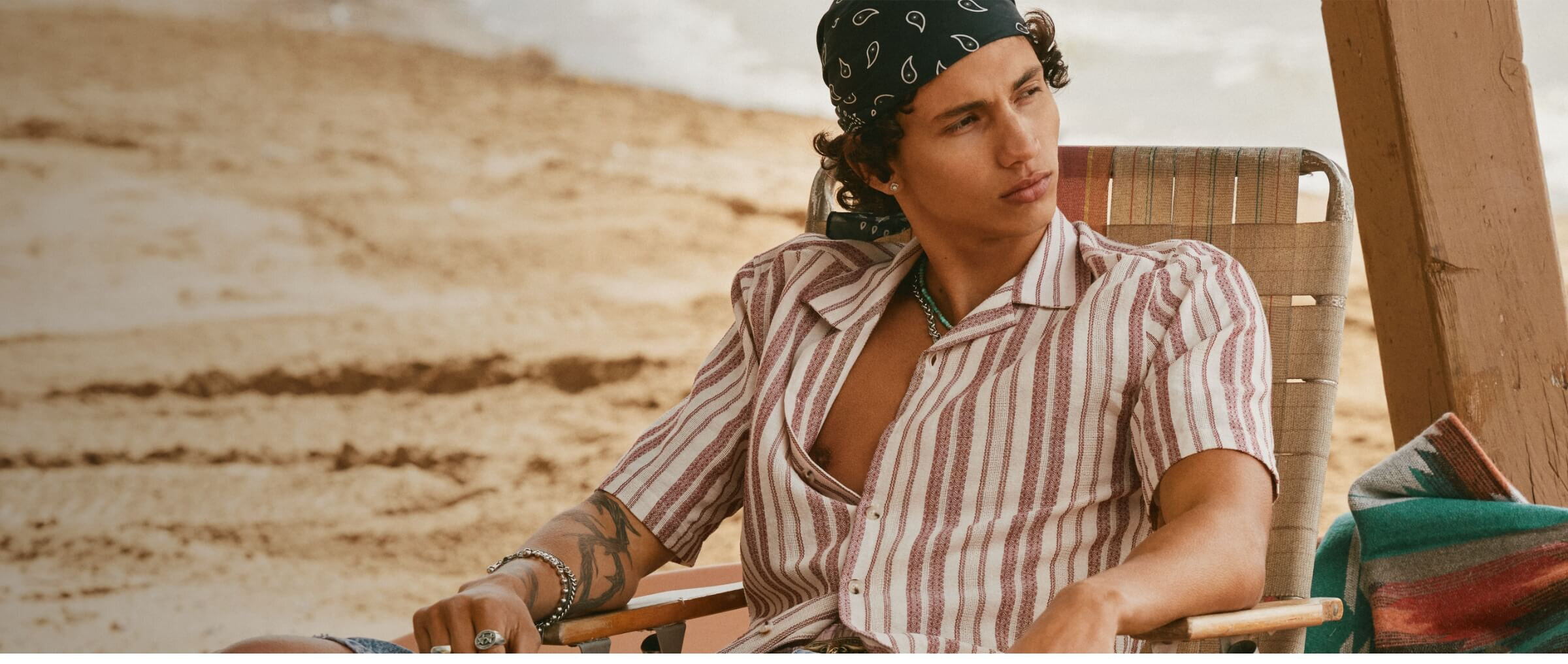 A young man in a cabana shirt and denim shorts sitting on a chair at the beach