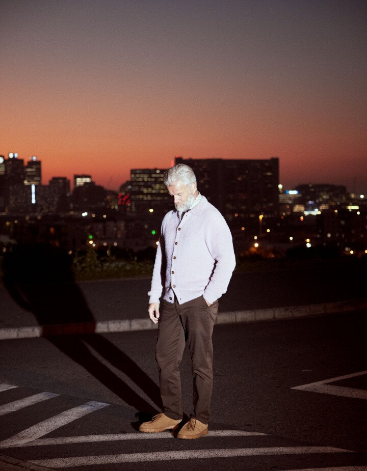 Elegant man with white hair and beard, wearing a lilac cardigan and brown pants, standing in a street overlooking the city at night