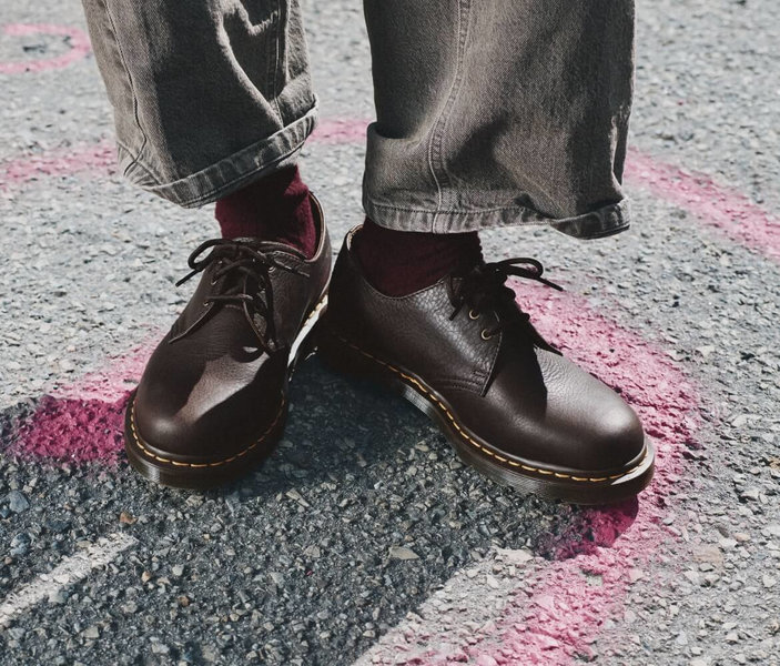 Gros plan sur des chaussures lacées en cuir brun foncé, portées avec un jean gris à revers et des chaussettes bordeaux.