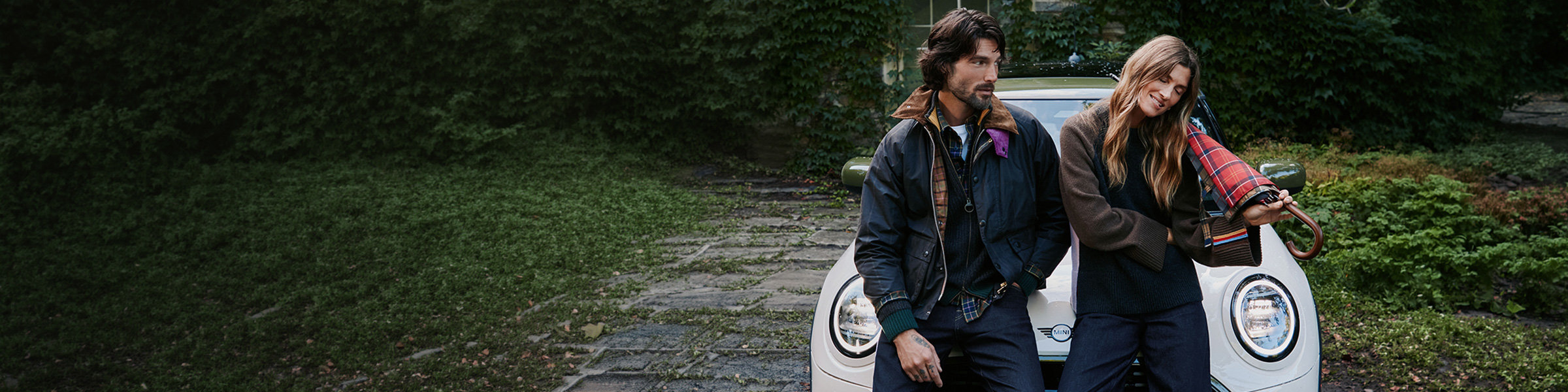 A man and a woman are sitting on the hood of a Mini Cooper. They are wearing jackets from the Barbour and Paul Smith collaboration. The woman is holding a tartan-patterned umbrella.