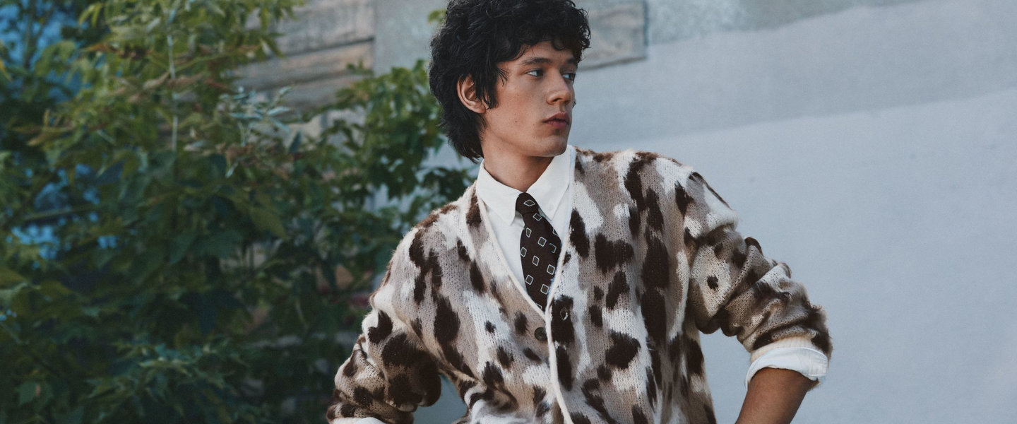 A man wears a brown and cream patterned cardigan over a white shirt and patterned tie.