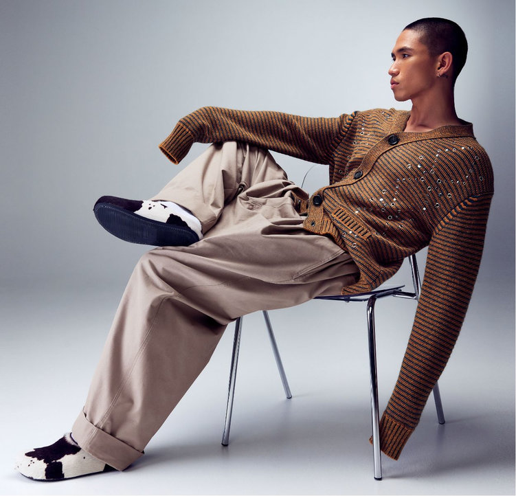 Jeune homme assis sur une chaise en position nonchalante, portant un cardigan rayé, un pantalon beige large et des mules motif vache de la marque Marni.