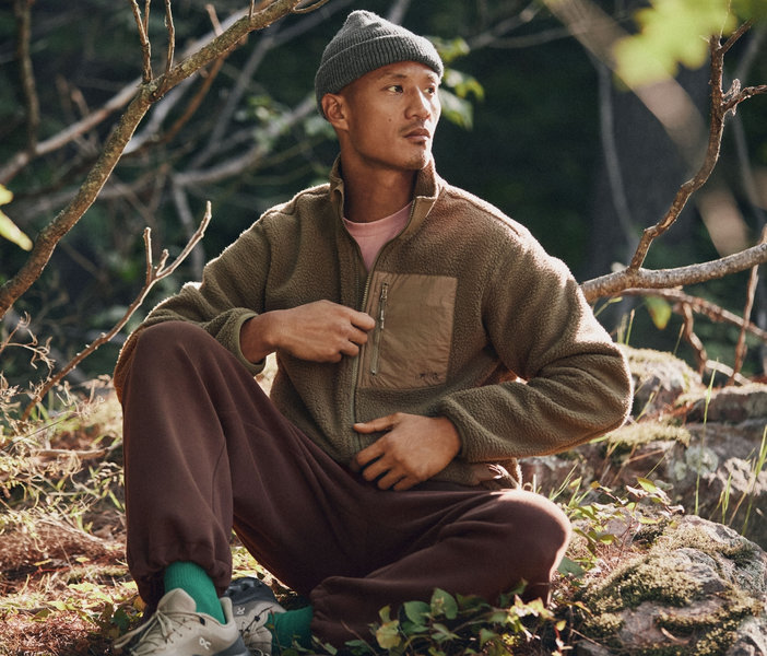 Man sitting on the forest floor, wearing a brown fleece jacket, dark pants, and a grey beanie.