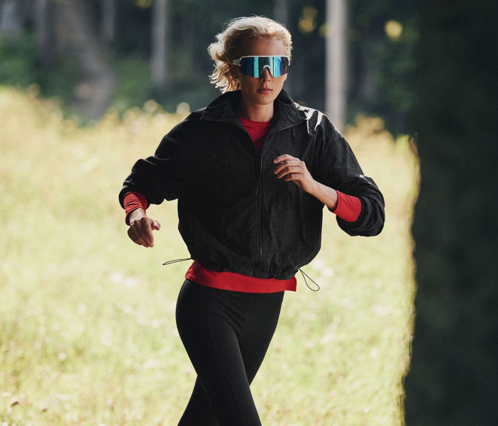 A woman runs through a forest wearing a black windbreaker, red top, black leggings, and mirrored sport sunglasses.
