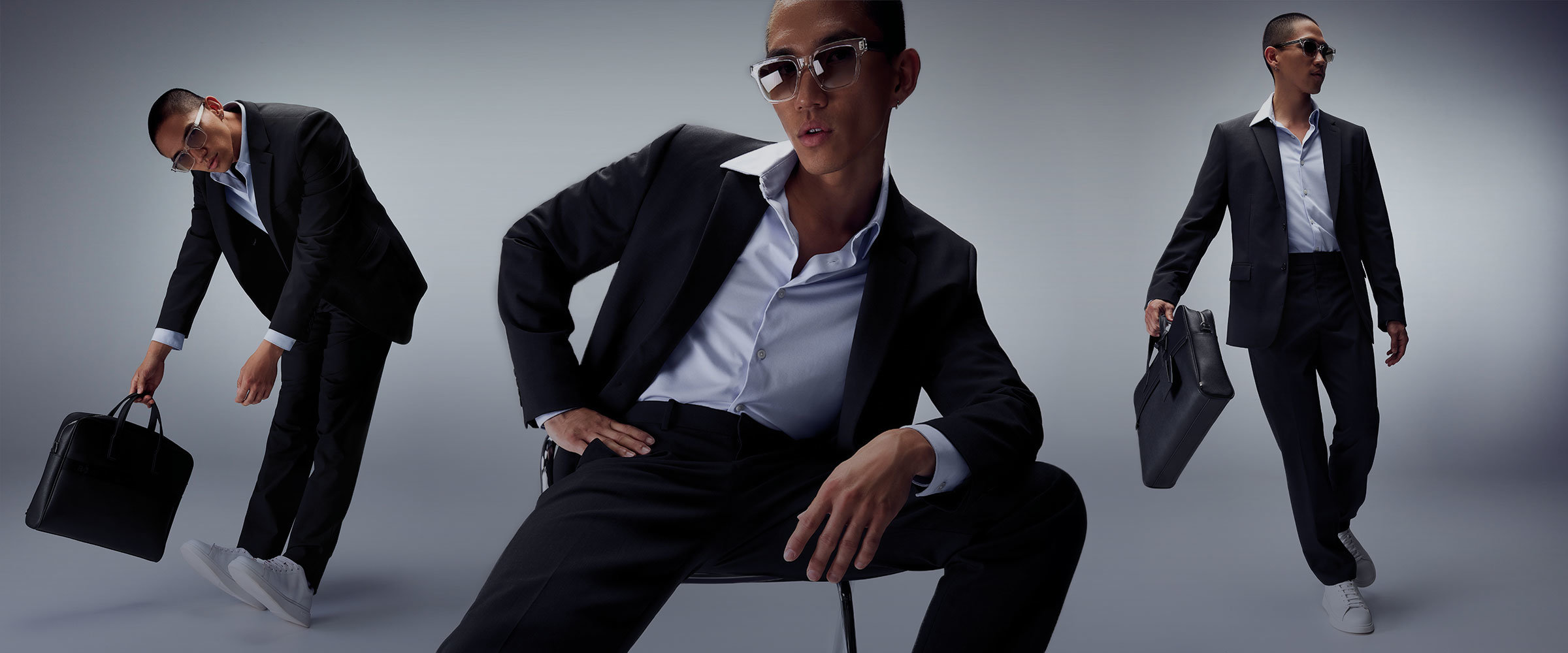 A young man striking a dynamic pose, wearing sunglasses and a dark suit with the jacket open to reveal a light-colored shirt. He is holding a briefcase.