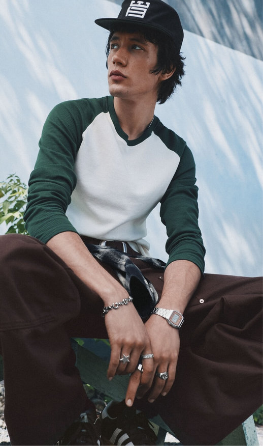 Seated man wearing a white-and-green baseball shirt, brown trousers and a black cap.