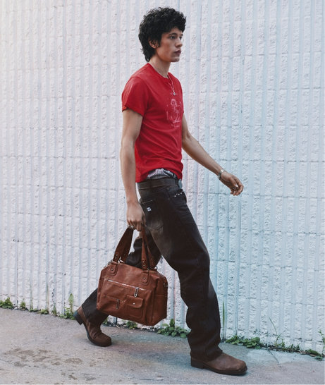 Homme marchant avec un sac brun, vêtu d'un t-shirt rouge, d'un jean foncé et de bottes brunes.
