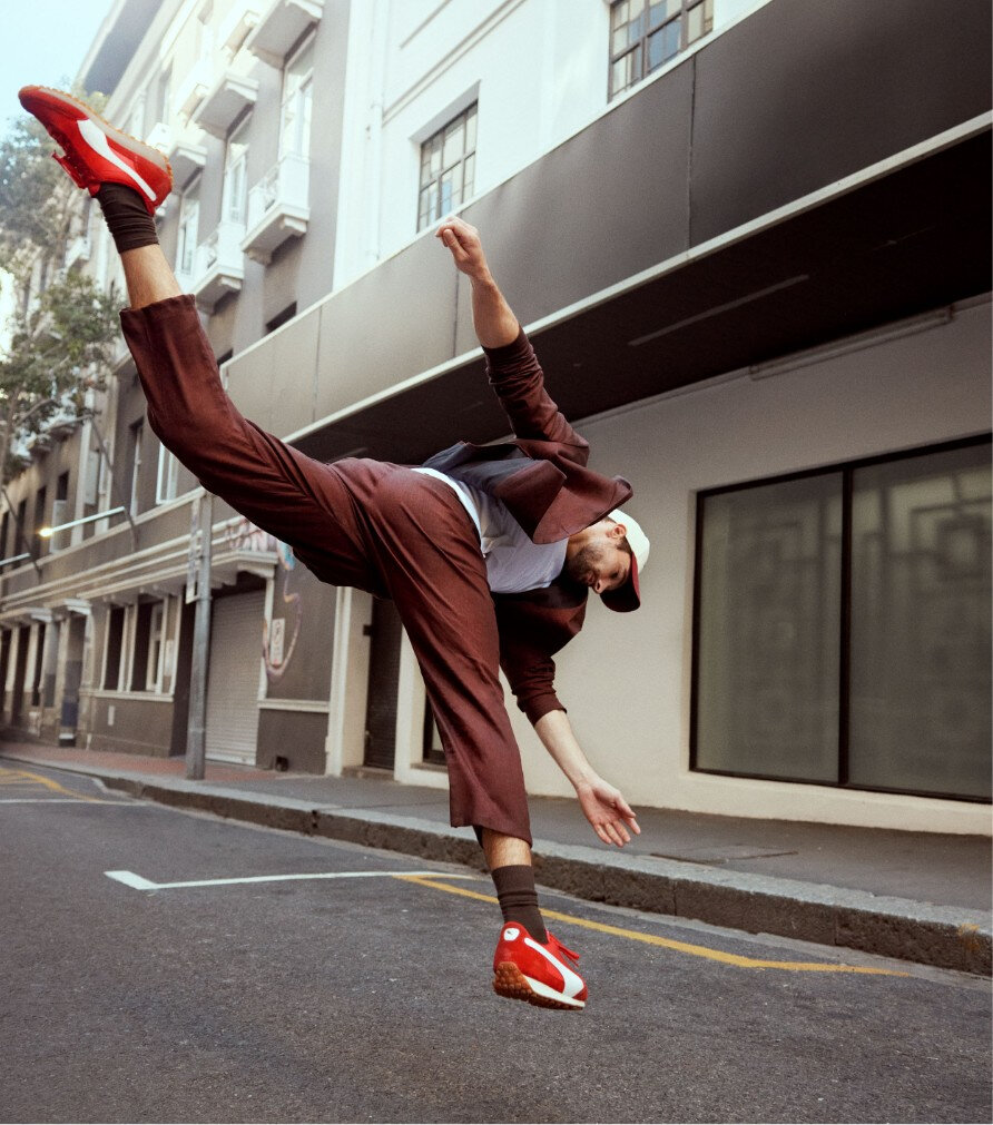 Homme en plein saut dans la rue, vêtu d'un complet bourgogne, t-shirt blanc, casquette et sneakers Puma rouges