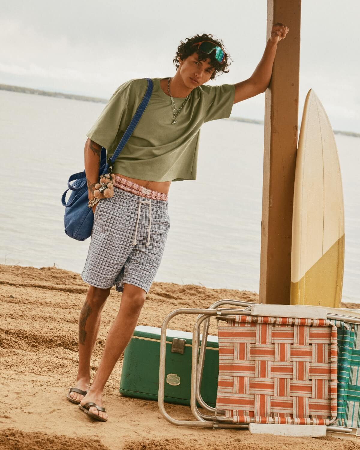 A young man at the beach wearing a loose-fitting, cropped khaki T-shirt and plaid shorts