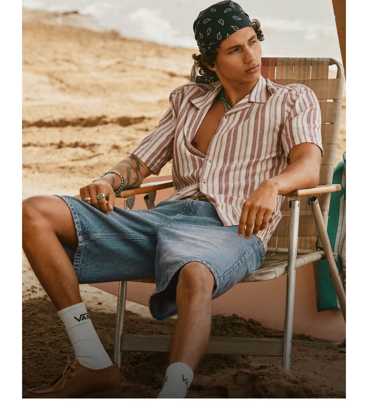 A young man in a cabana shirt and denim shorts sitting on a chair at the beach