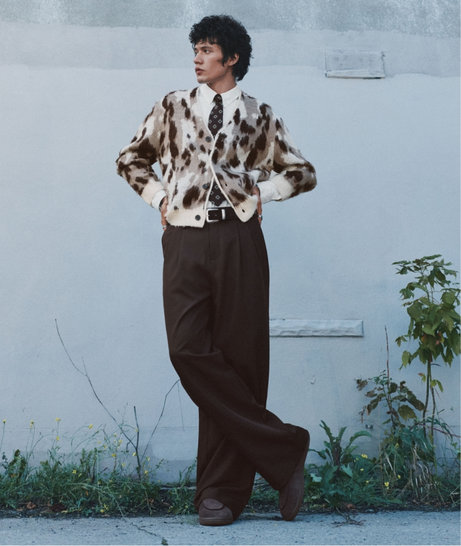 Man standing against a pale wall in a brown-and-cream patterned cardigan and wide-leg brown trousers.