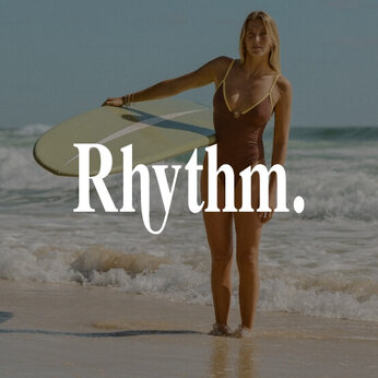 Une femme sur la plage avec une planche de surf porte un maillot une-pièce brun et jaune de marque Rhythm.