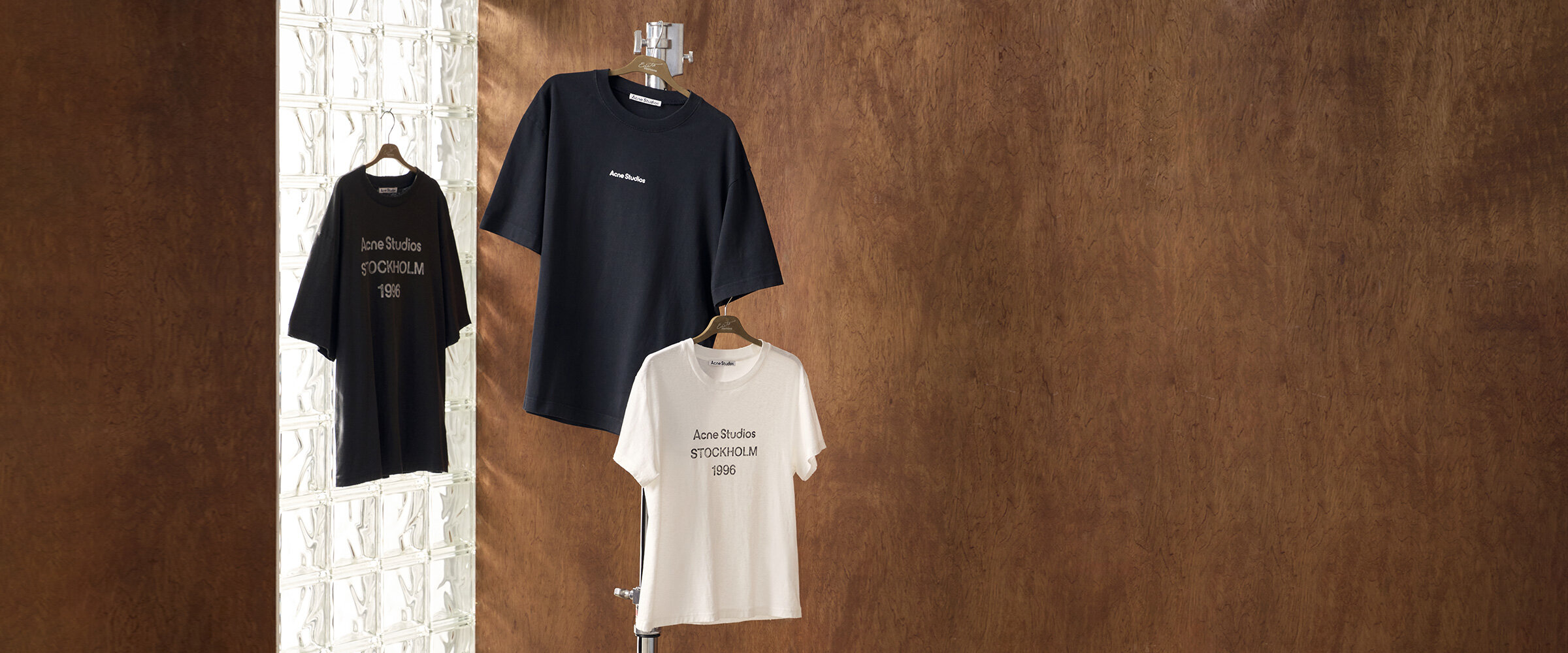 Des t-shirts de la marque Acne Studios sont suspendus sur des cintres sur un fond boisé.