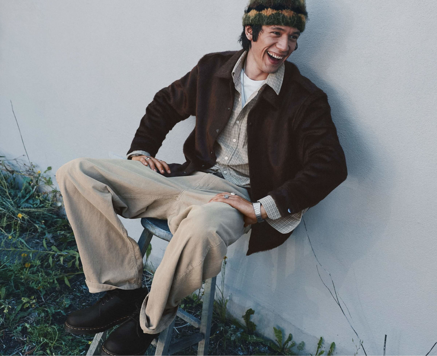 Man laughing on a chair, wearing a dark brown jacket, plaid shirt, beige trousers and striped beanie.